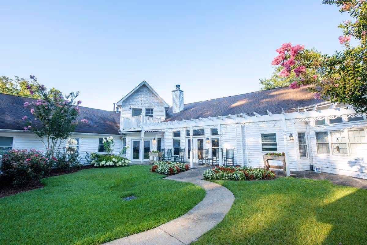 Exterior view of a white senior living facility building with a brown roof, surrounded by a well-maintained green lawn and colorful flower beds. There is a curved concrete pathway leading to a patio area with rocking chairs under a white pergola. Trees with pink flowers frame the scene under a clear blue sky.