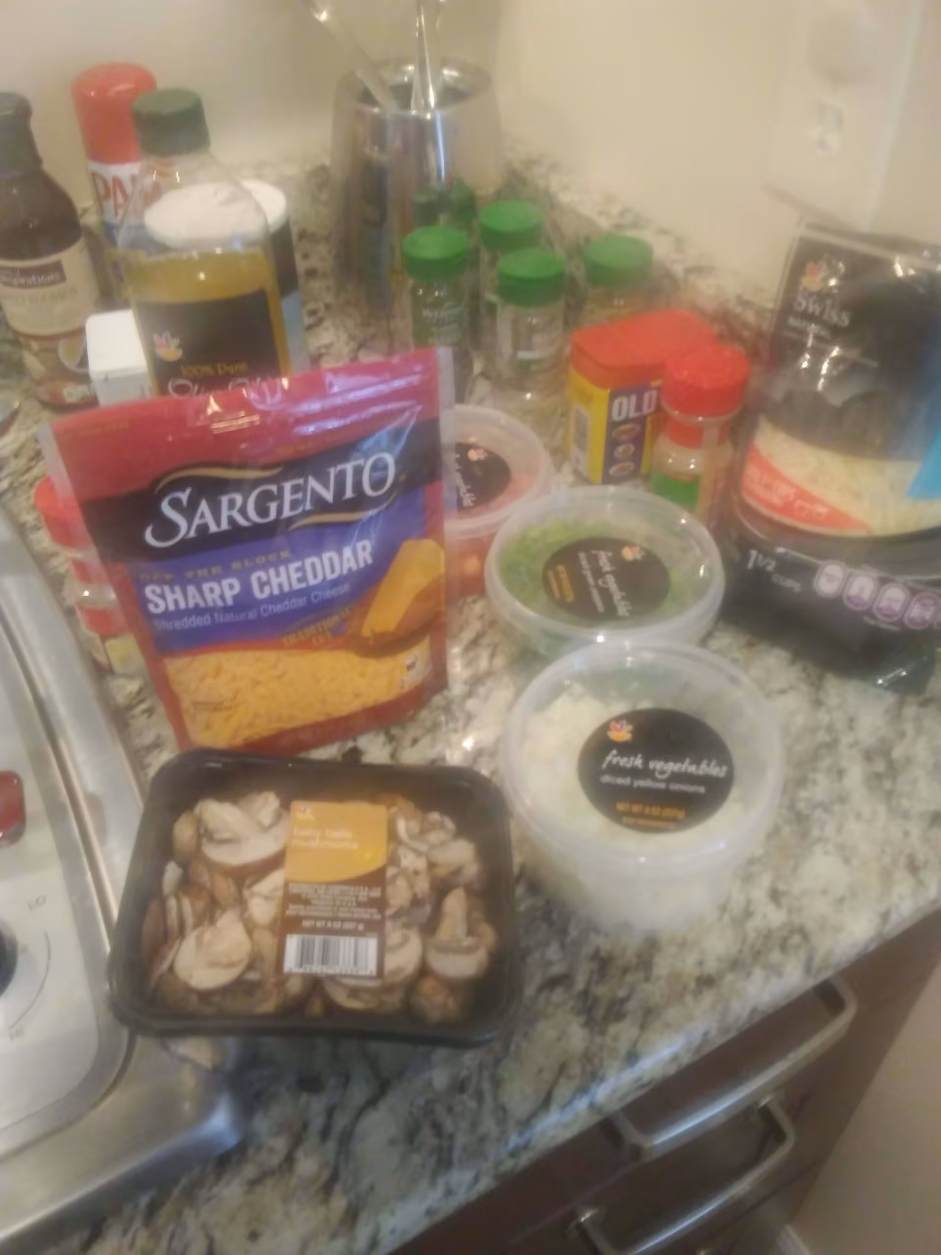 A kitchen countertop with various food items including a bag of Sargento sharp cheddar shredded cheese, containers of fresh vegetables like diced onions and chopped green peppers, a package of sliced mushrooms, several spice bottles, and other cooking ingredients.