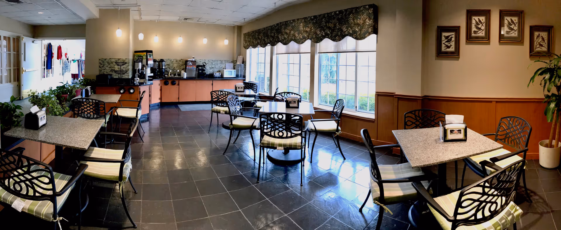 A bright dining area with several tables and chairs arranged neatly. The room features large windows with patterned valances allowing natural light to fill the space. There is a counter area at the back with coffee machines and other appliances. The walls are decorated with framed artwork and a potted plant is visible in the corner.