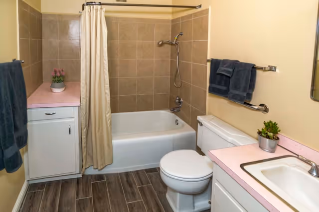 A clean bathroom with a bathtub and shower, toilet, sink with pink countertops, cabinets, towels, and small potted plants.