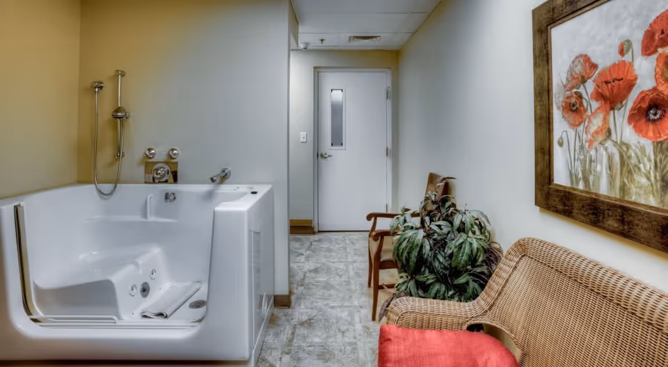 A small room with a white walk-in bathtub on the left side, equipped with a handheld showerhead and faucet. On the right side, there is a wicker bench with a red cushion, a potted plant, and a wooden chair. A framed painting of red poppies hangs on the wall above the bench. At the end of the room, there is a white door with a small window.