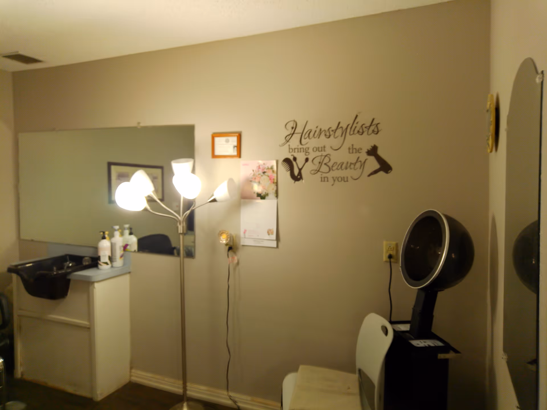 Interior of a hair salon area with a black hair washing sink, a large mirror, a standing lamp with multiple lights, a calendar on the wall, a chair, and a hair dryer hood. The wall has a decorative text that reads 'Hairstylists bring out the Beauty in you'.