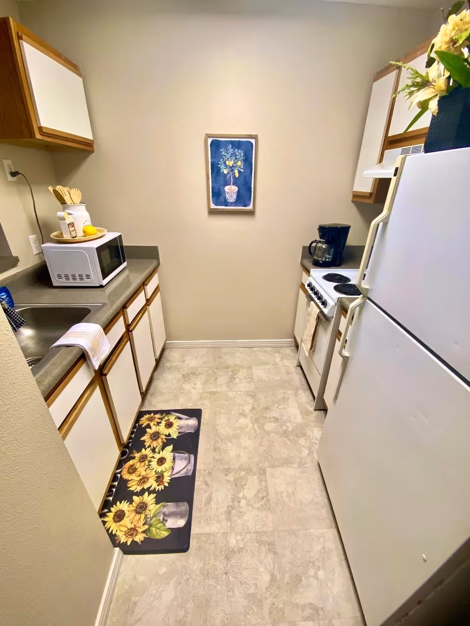 A small kitchen with white cabinets and wooden trim, a white refrigerator, a white stove with a coffee maker on the counter, a microwave on the opposite counter, a sink with a towel hanging over the edge, a sunflower-themed floor mat, and a framed picture of a potted plant on the wall.