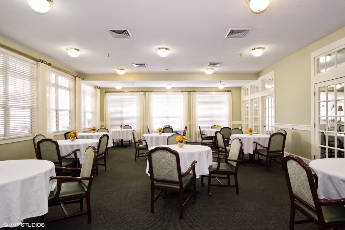 Bright dining room with round tables covered in white tablecloths and chairs, each table decorated with a small floral centerpiece.