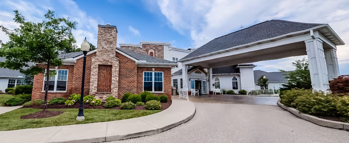 Front exterior view of Birchwood of Hamilton facility showing a brick building with a stone chimney, a covered entrance with white pillars, landscaped greenery, and a clear sky with some clouds.