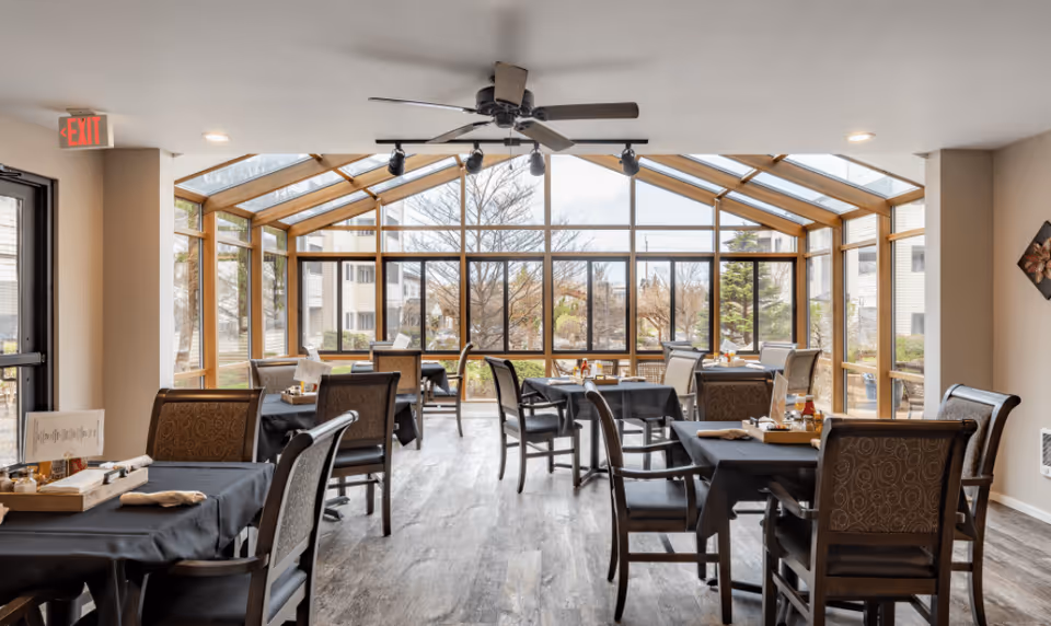 Bright glass-enclosed dining area with multiple set tables and chairs facing a courtyard view.