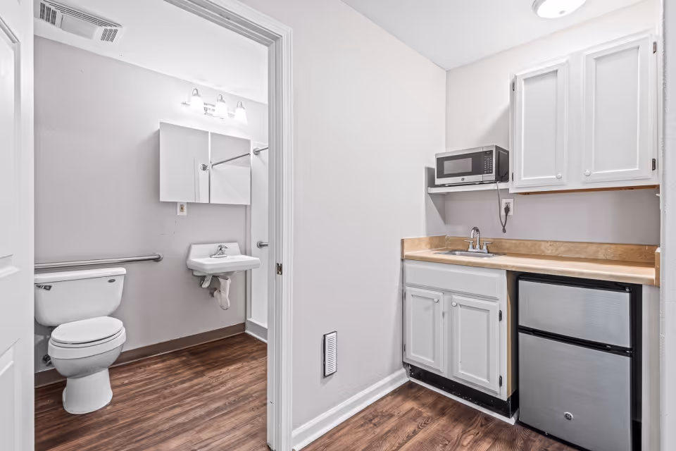 A small kitchenette area with white cabinets, a countertop with a sink, a microwave on a shelf above, and a mini refrigerator below. To the left, an open door reveals a bathroom with a toilet, a wall-mounted sink, a mirror cabinet, and a shower with a grab bar. The floor is wood-style laminate, and the walls are painted white.