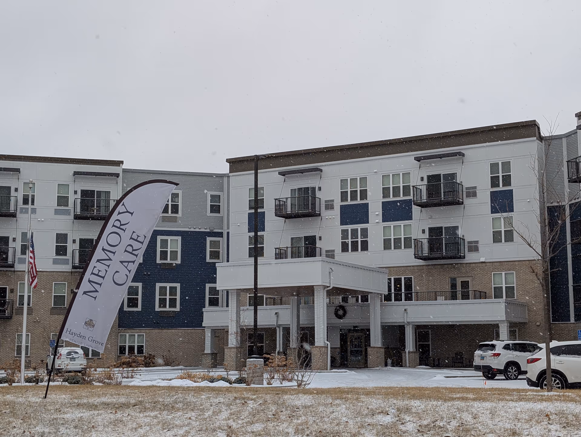 Snowy front exterior of a multi-story senior living building with balconies and a 'Memory Care' flag by the entrance.