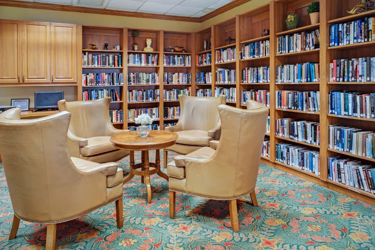 A cozy library room with four beige leather armchairs arranged around a small round wooden table with a glass vase holding white flowers. The walls are lined with wooden bookshelves filled with books, and there are wooden cabinets and a small computer on a desk in the corner. The floor is covered with a patterned carpet in shades of green, red, and yellow.