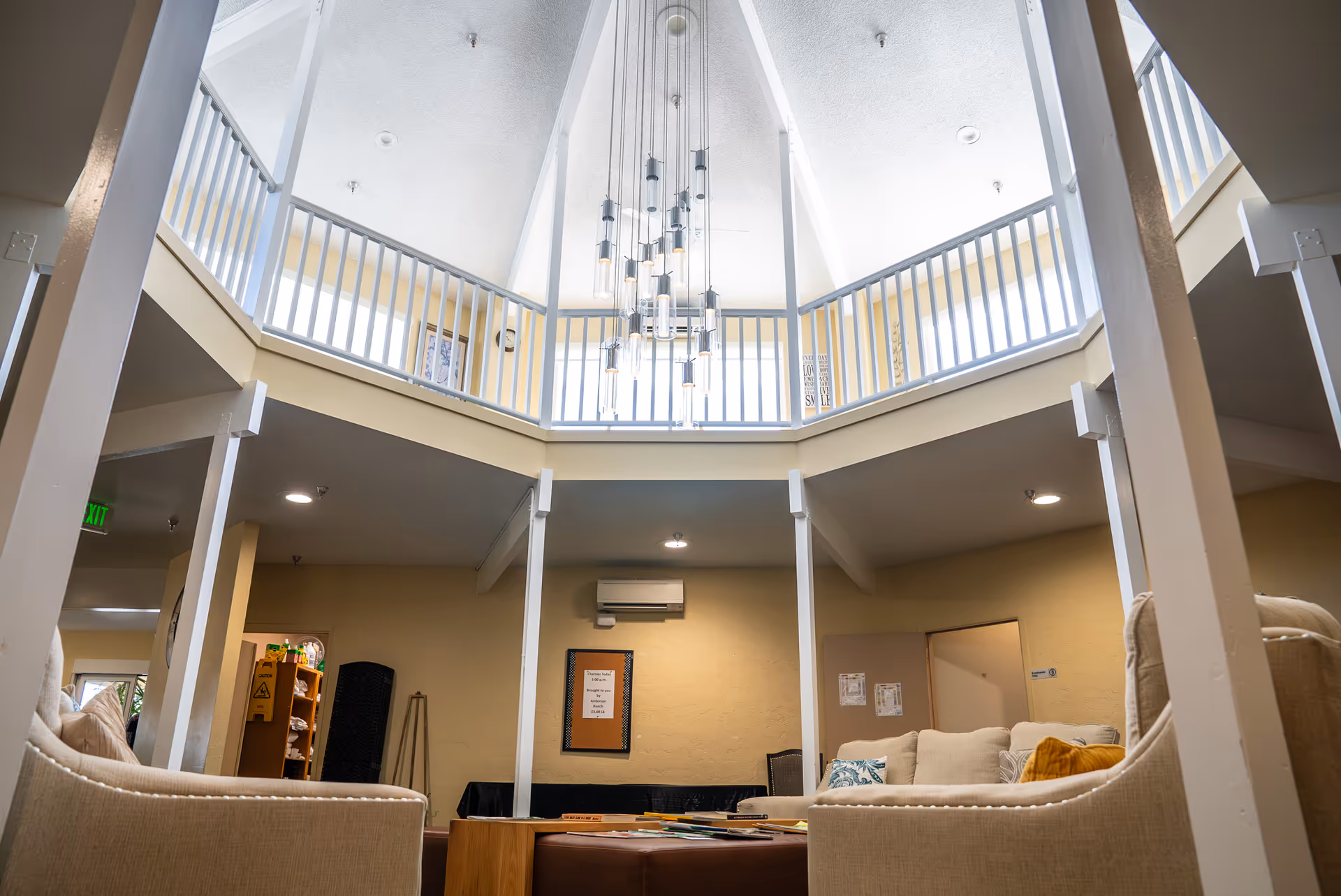 Interior view of a senior living facility common area with beige walls and high vaulted ceiling. The room features a modern hanging light fixture, white railings on the upper level, beige cushioned chairs, and a bulletin board on the wall. There are some books and magazines on a central table.