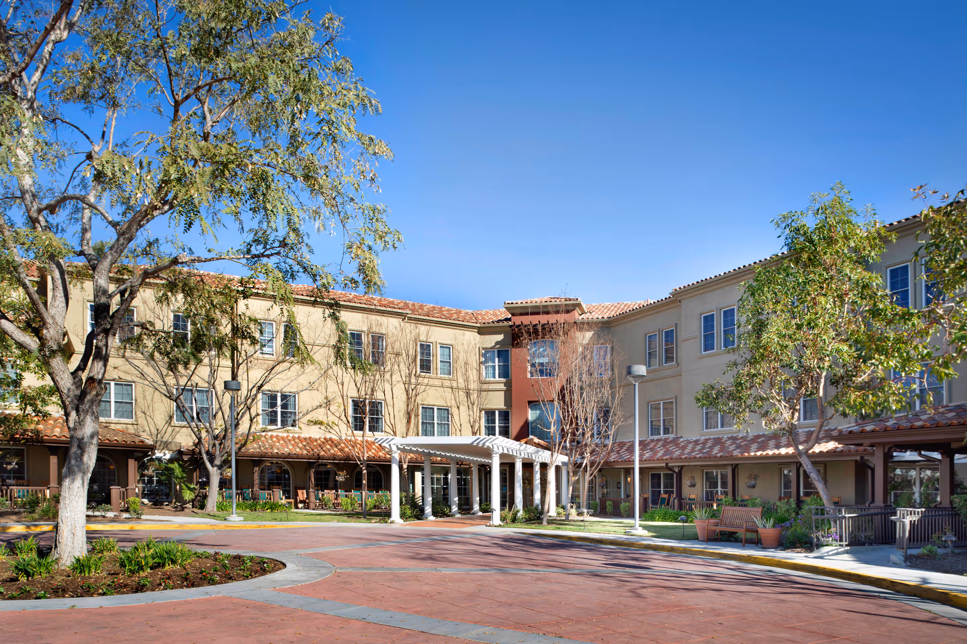 Exterior view of a three-story senior living facility building with beige and brown walls, multiple windows, a covered entrance with white columns, trees, and a clear blue sky.