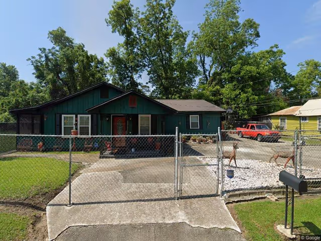 Front view of a single-story green house with a chain-link fence, driveway, and a red pickup truck parked to the side.
