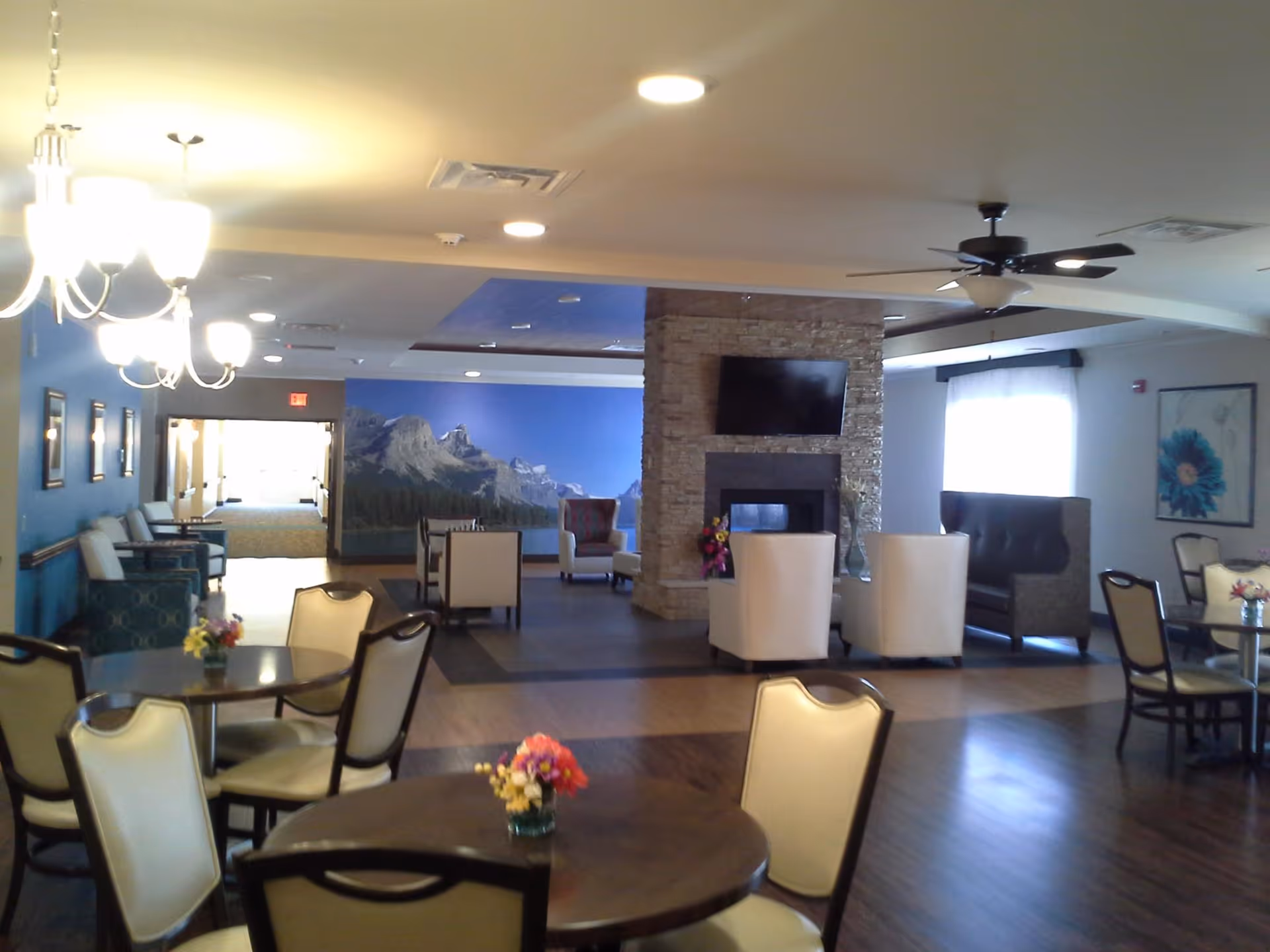 Open, well-lit senior common area with round dining tables and chairs in the foreground and a stone fireplace with seating and a TV in the background.