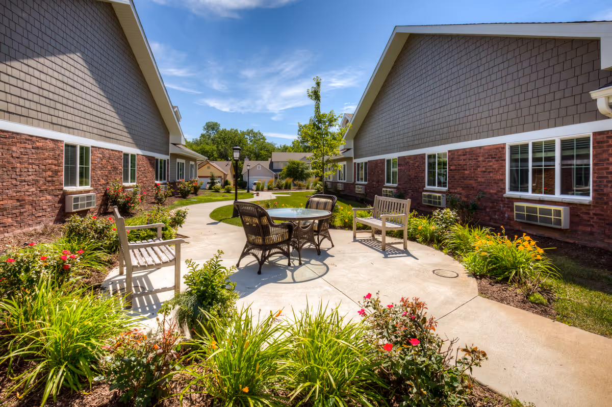 Sunny courtyard between two single-story brick residential buildings with a round patio table, chairs and benches surrounded by landscaped flower beds.