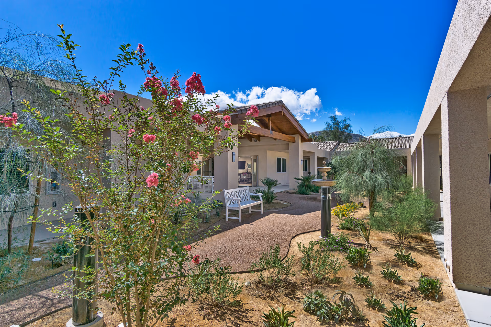 Outdoor courtyard area of a senior living facility with a clear blue sky, flowering plants, shrubs, a white bench, a water fountain, and beige buildings with tiled roofs surrounding the garden.