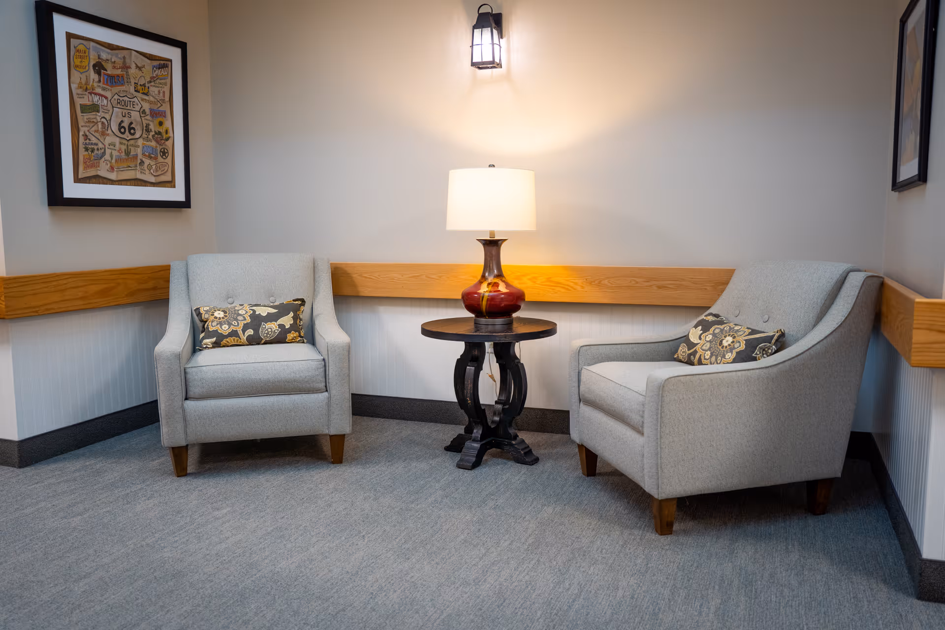 Two gray upholstered armchairs with patterned pillows flank a small round table holding a lamp in a cozy lobby seating area.