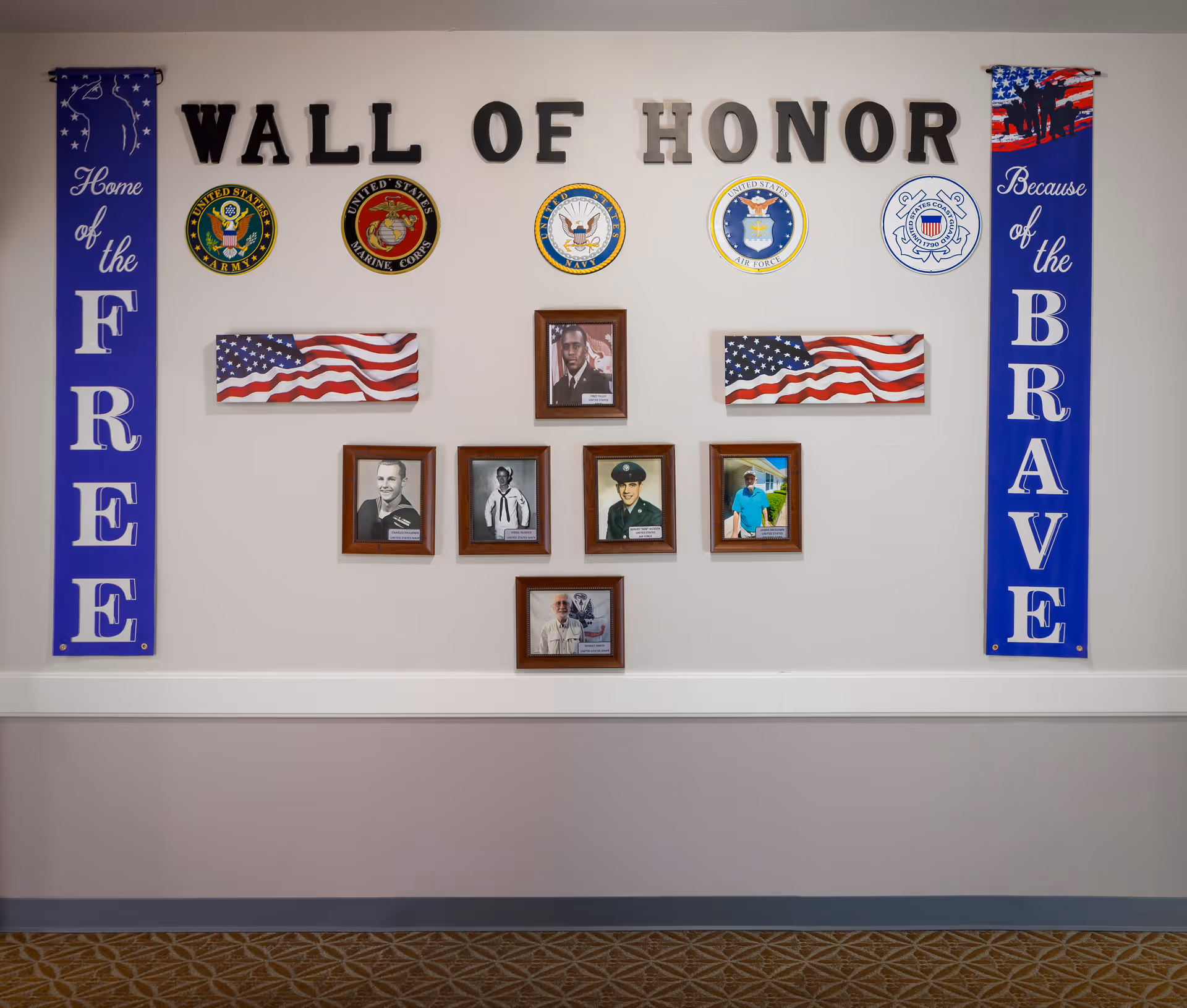 A Wall of Honor display featuring the emblems of the United States Army, Marine Corps, Navy, Air Force, and Coast Guard. The display includes the words 'WALL OF HONOR' in large black letters at the top, two American flag plaques, and six framed photographs of veterans. On the left side, a blue banner reads 'Home of the FREE' and on the right side, a blue banner reads 'Because of the BRAVE.'
