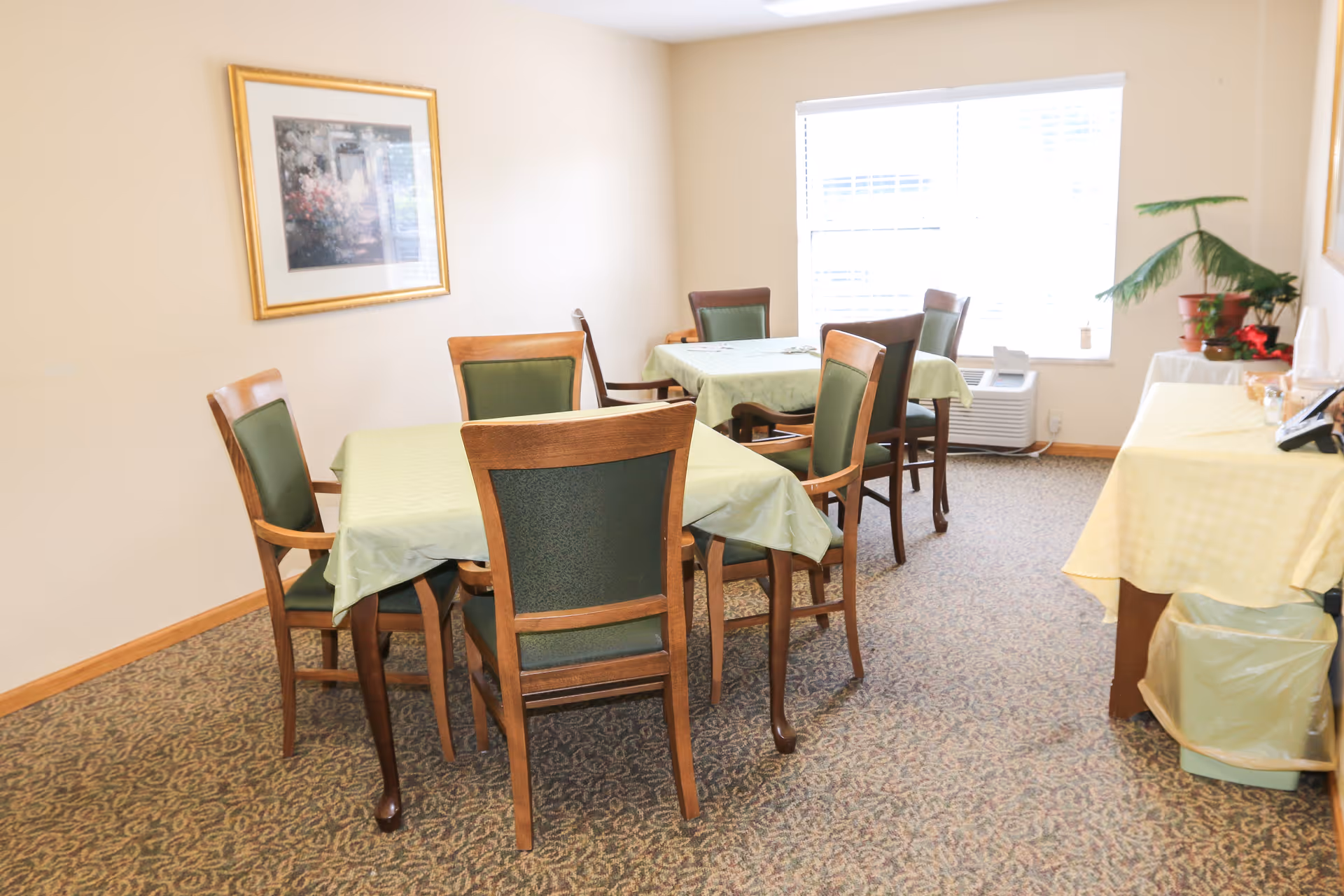A small dining room with two tables covered with light green tablecloths, each surrounded by four wooden chairs with green cushioned backs and seats. There is a framed picture on the beige wall, a window letting in natural light, and a side table with a yellow tablecloth holding some plants and other items. The floor is carpeted with a patterned design.