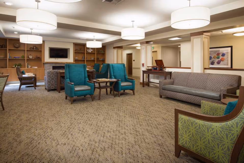 Spacious, well-lit senior living lounge with teal armchairs, a sofa, built-in shelves and pendant lights.