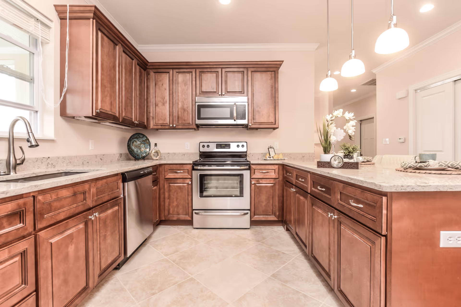 Bright kitchen with wooden cabinets, stainless steel appliances including a stove, microwave, and dishwasher, a double sink under a window, and a countertop with decorative items and pendant lights overhead.