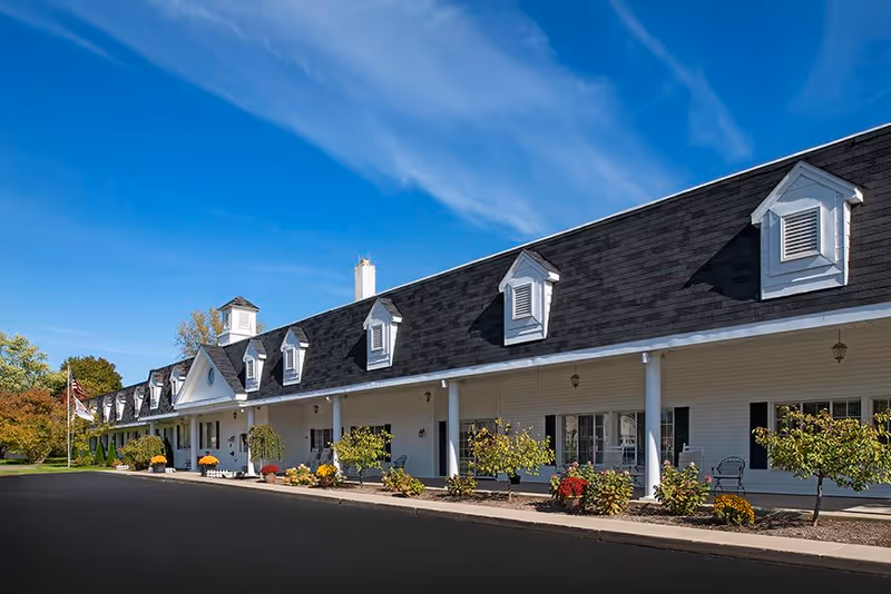 Exterior view of a long, white senior living facility building with a black shingled roof featuring multiple dormer windows. The building has a covered porch with white columns, landscaped bushes, and flowers along the front. The sky is clear and blue.