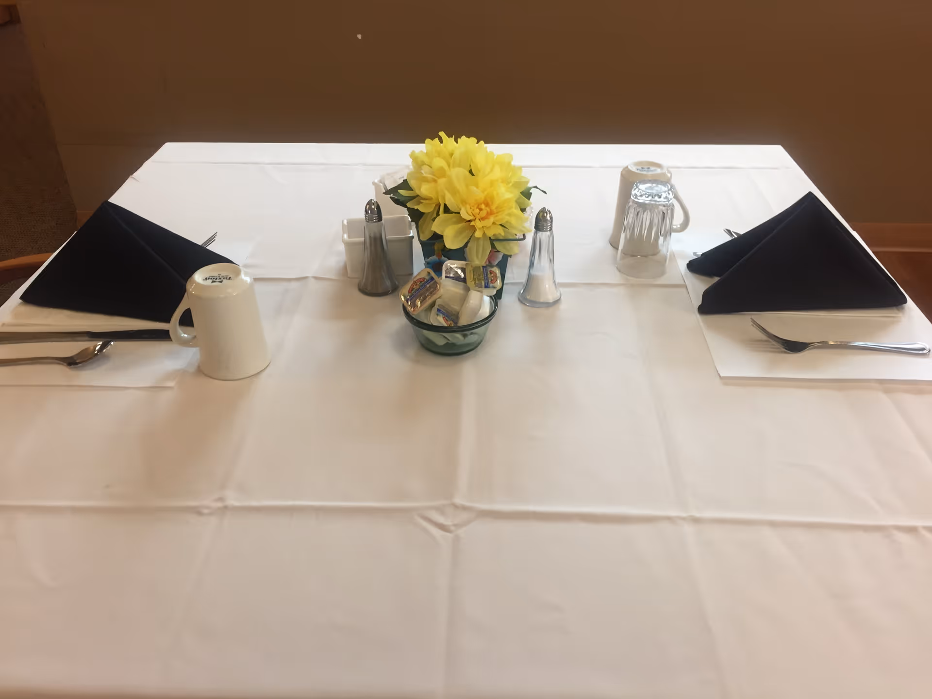 Table set for two with black napkins, mugs, silverware, salt and pepper, and a small vase of yellow flowers on a white tablecloth.