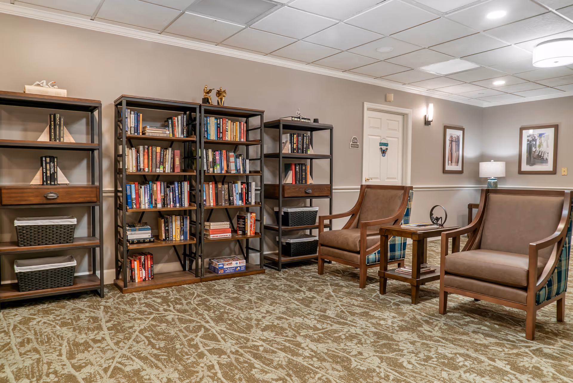 Cozy communal seating area with bookshelves, two armchairs and a side table in a senior living facility.