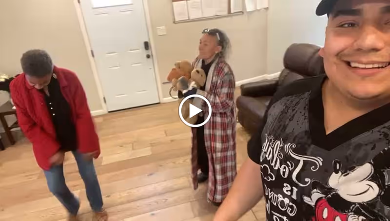 A smiling man taking a selfie in a living room with two women in the background. One woman is wearing a red jacket and looking down, while the other is wearing a plaid robe and holding two teddy bears. The room has wooden flooring, a white door, and a brown leather chair.