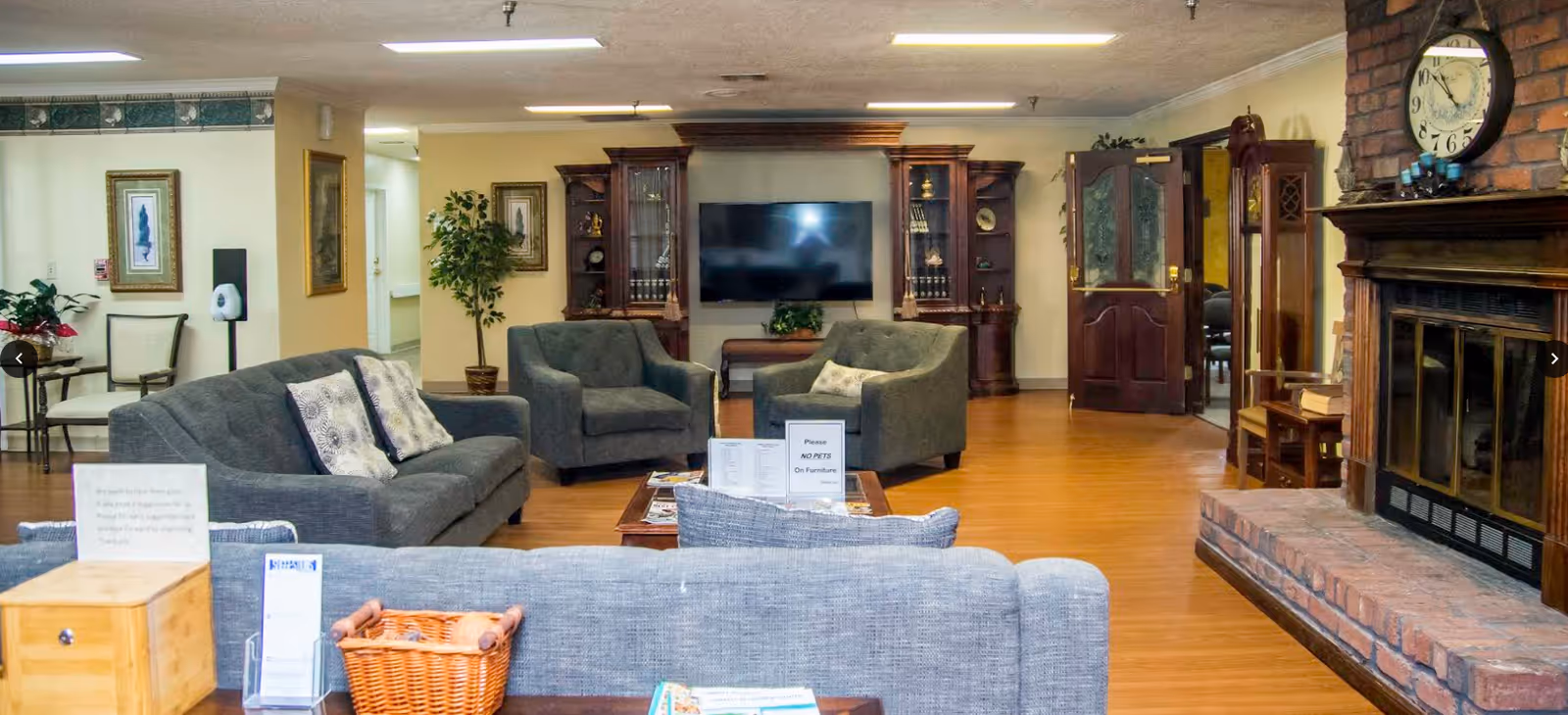 A cozy living room area in a retirement center featuring several gray upholstered sofas and armchairs arranged around a wooden coffee table with signs and magazines. The room has a large flat-screen TV mounted on the wall between two wooden display cabinets. There is a brick fireplace with a clock above it on the right side, wooden flooring, framed artwork on the walls, and a potted plant near the hallway entrance.