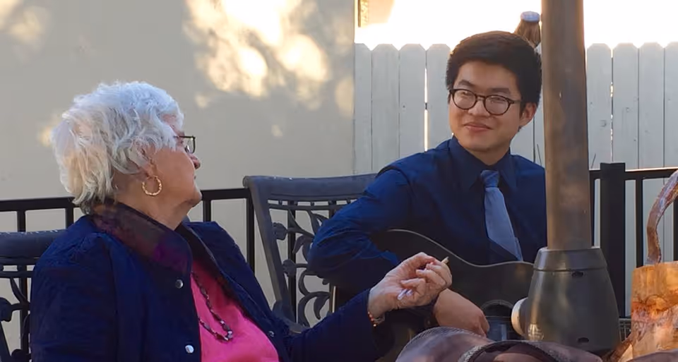 An elderly woman with white hair and glasses sitting outdoors, holding hands with a young man wearing glasses, a blue shirt, and a tie who is playing a guitar. They are seated on metal chairs near a patio heater with a white fence in the background.