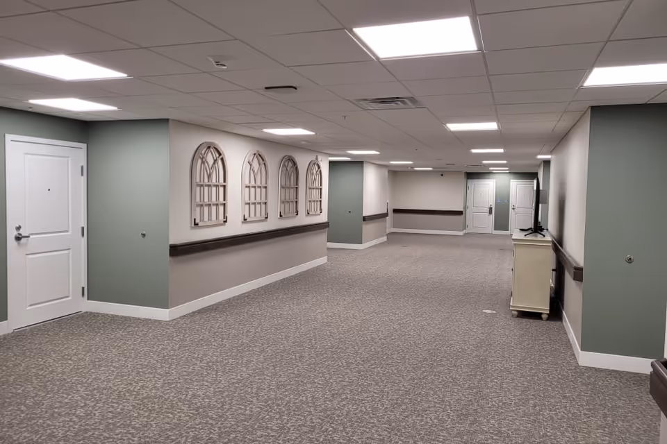 A wide, carpeted hallway in a senior living facility with light green and beige walls, white doors, and decorative wall hangings shaped like window frames. The ceiling has recessed lighting panels, and there is a small cabinet with a TV on the right side.
