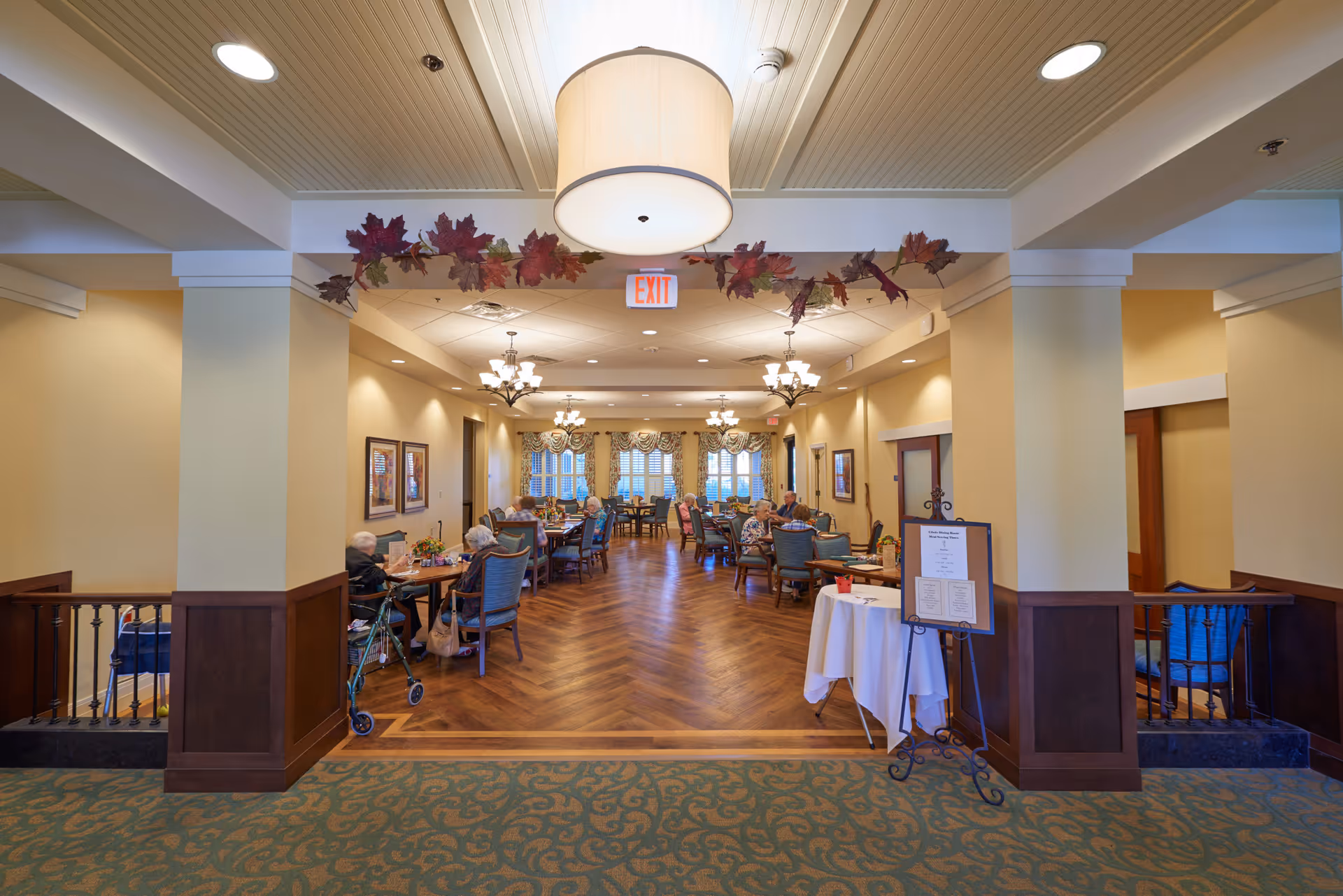 Dining room interior of a senior living facility with tables and seated residents under chandeliers and fall decorations.