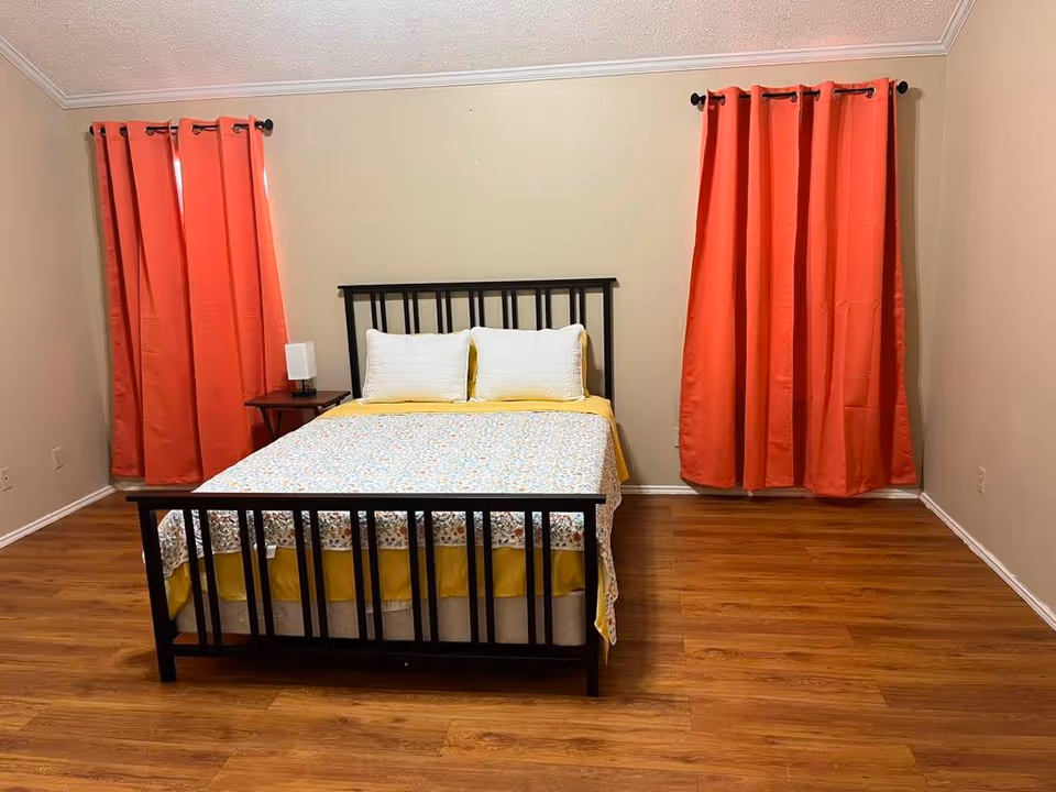 Simple bedroom with a black metal bed centered between two windows with red curtains, wood floors, and a small nightstand with a lamp.