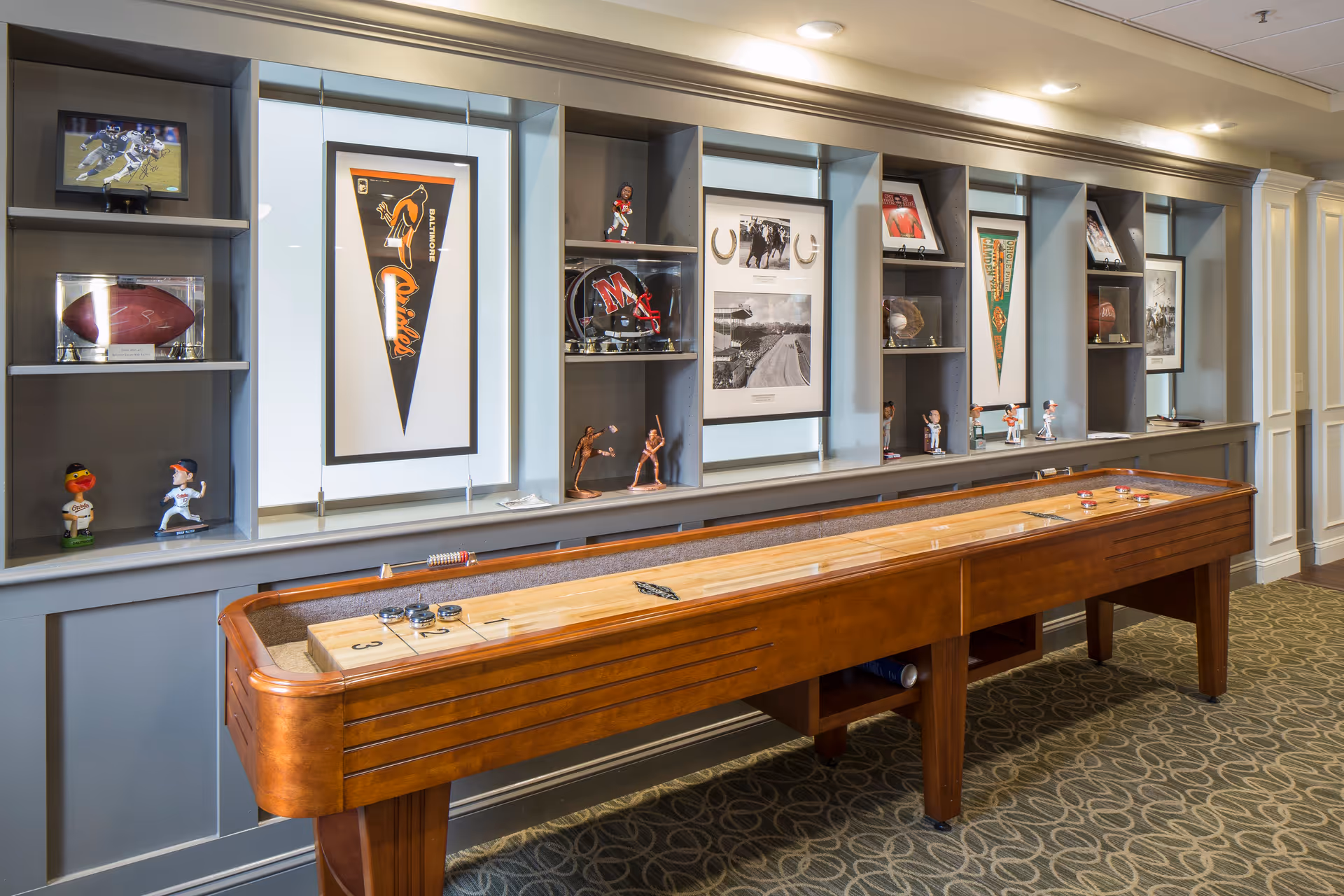 A game room with a wooden shuffleboard table in the foreground and a built-in wall shelf in the background displaying sports memorabilia including framed pennants, helmets, bobblehead figures, and photographs.