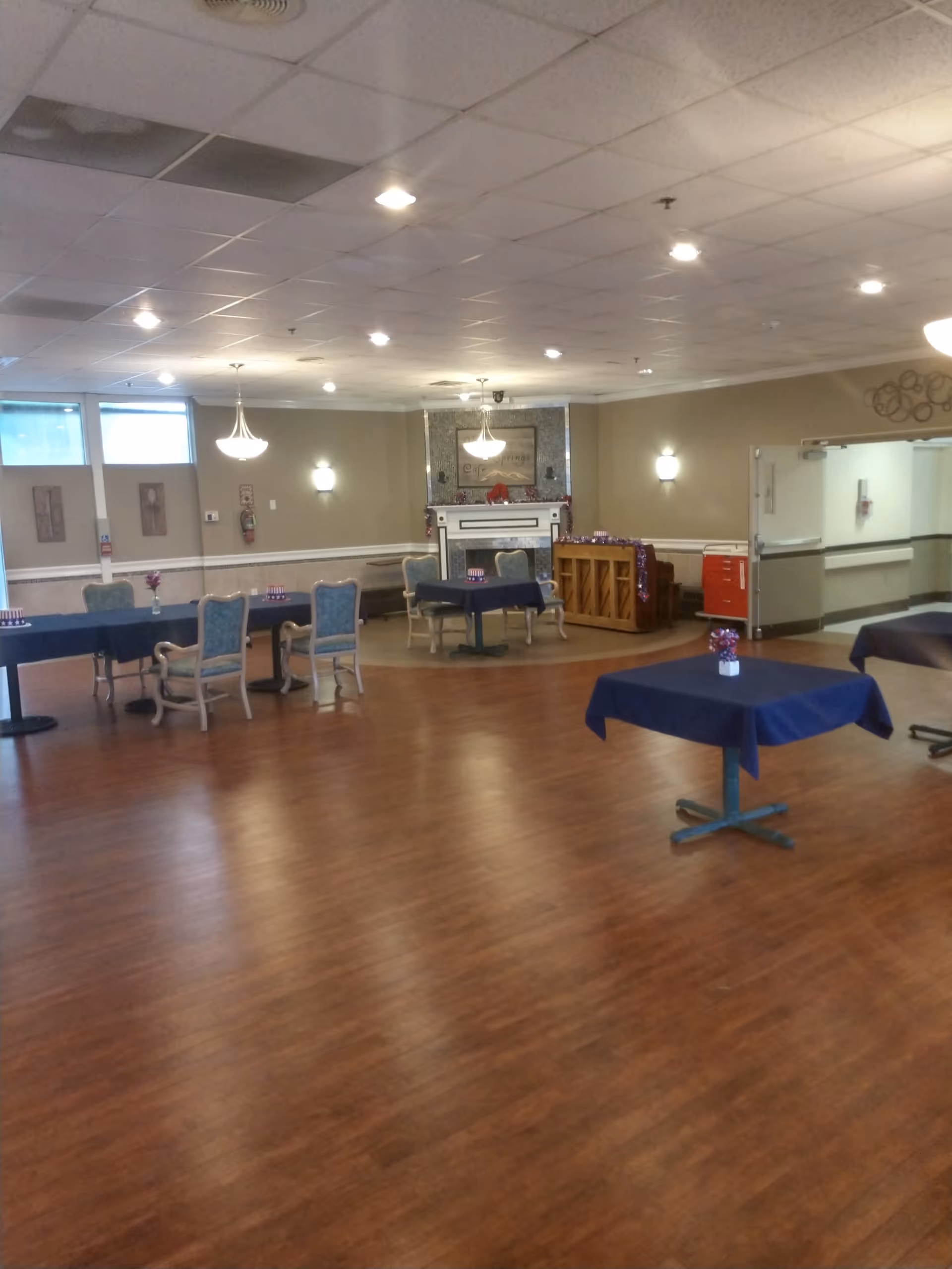 A spacious common area with wooden flooring, several tables covered with dark blue tablecloths, and chairs arranged around them. The room features a fireplace with a decorative mantle, a piano, and soft lighting from ceiling fixtures and wall sconces. The walls are painted beige with white trim, and there are windows letting in natural light.