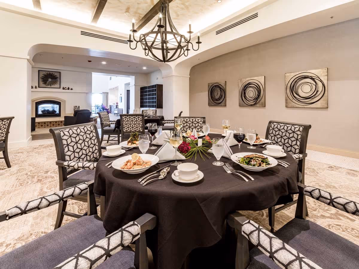 A dining room with a round table covered with a black tablecloth set for a meal with plates of food, glasses of water and wine, and silverware. The room has patterned chairs around the table, a chandelier hanging from the ceiling, abstract circular artwork on the wall, and a fireplace in the background with additional seating.