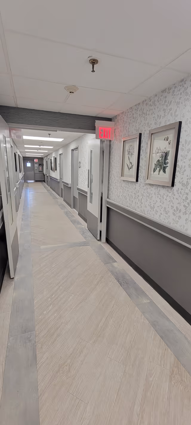 Long, well-lit interior corridor in a senior living facility with handrails, framed botanical prints, patterned wallpaper and an illuminated EXIT sign.