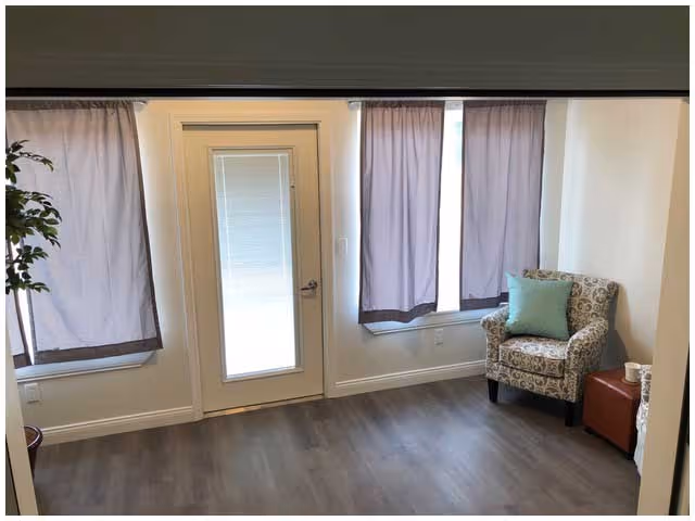 A small sitting area with a patterned armchair, a green cushion, a small brown ottoman with a cup on it, and a potted plant. The room has light-colored walls, wood flooring, two windows with purple curtains, and a door with a window covered by a blind.