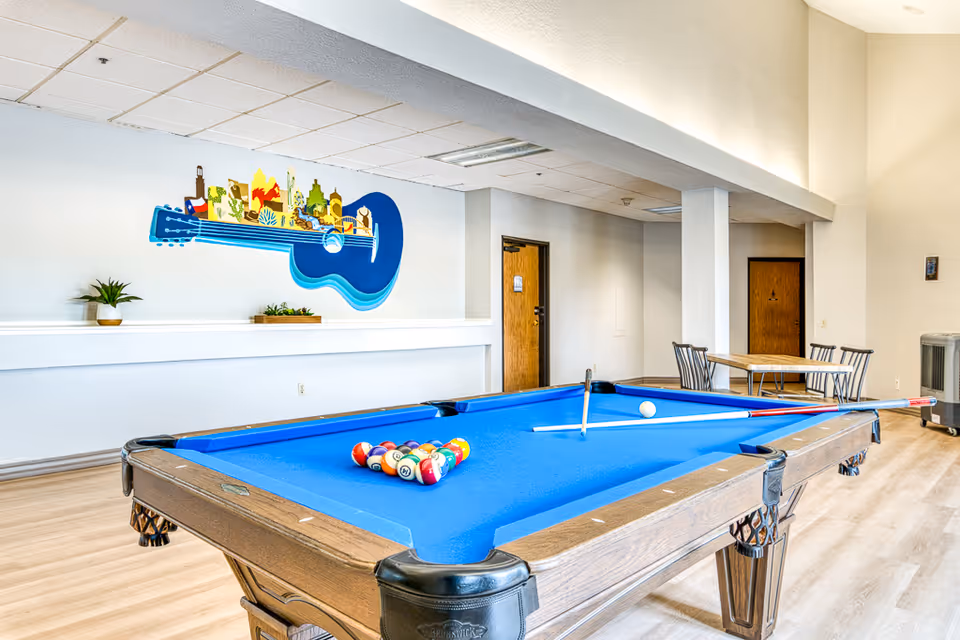 A bright recreational room featuring a blue felt pool table with billiard balls arranged for a game. The room has light wood flooring, white walls, and a colorful wall decoration shaped like a guitar with various cityscape elements. In the background, there is a small table with four chairs and two wooden doors.