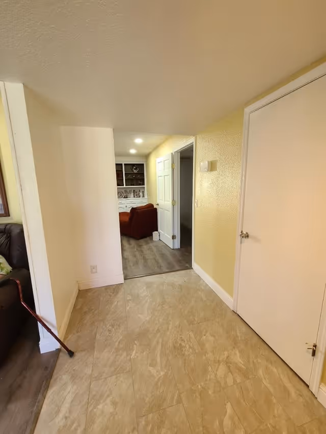 Interior view of a hallway with beige tiled floor and yellow walls leading to a room with wooden flooring and a red armchair. There is a white door on the right side and a walking cane leaning against a brown couch partially visible on the left.