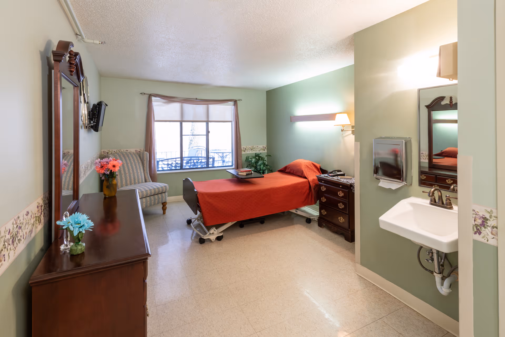 A well-lit room in a senior living facility featuring a single bed with a red bedspread, a wooden dresser with a mirror, a small armchair near a window with curtains, a wall-mounted television, and a sink with a mirror and paper towel dispenser on the right wall. The walls are painted light green with floral wallpaper border.