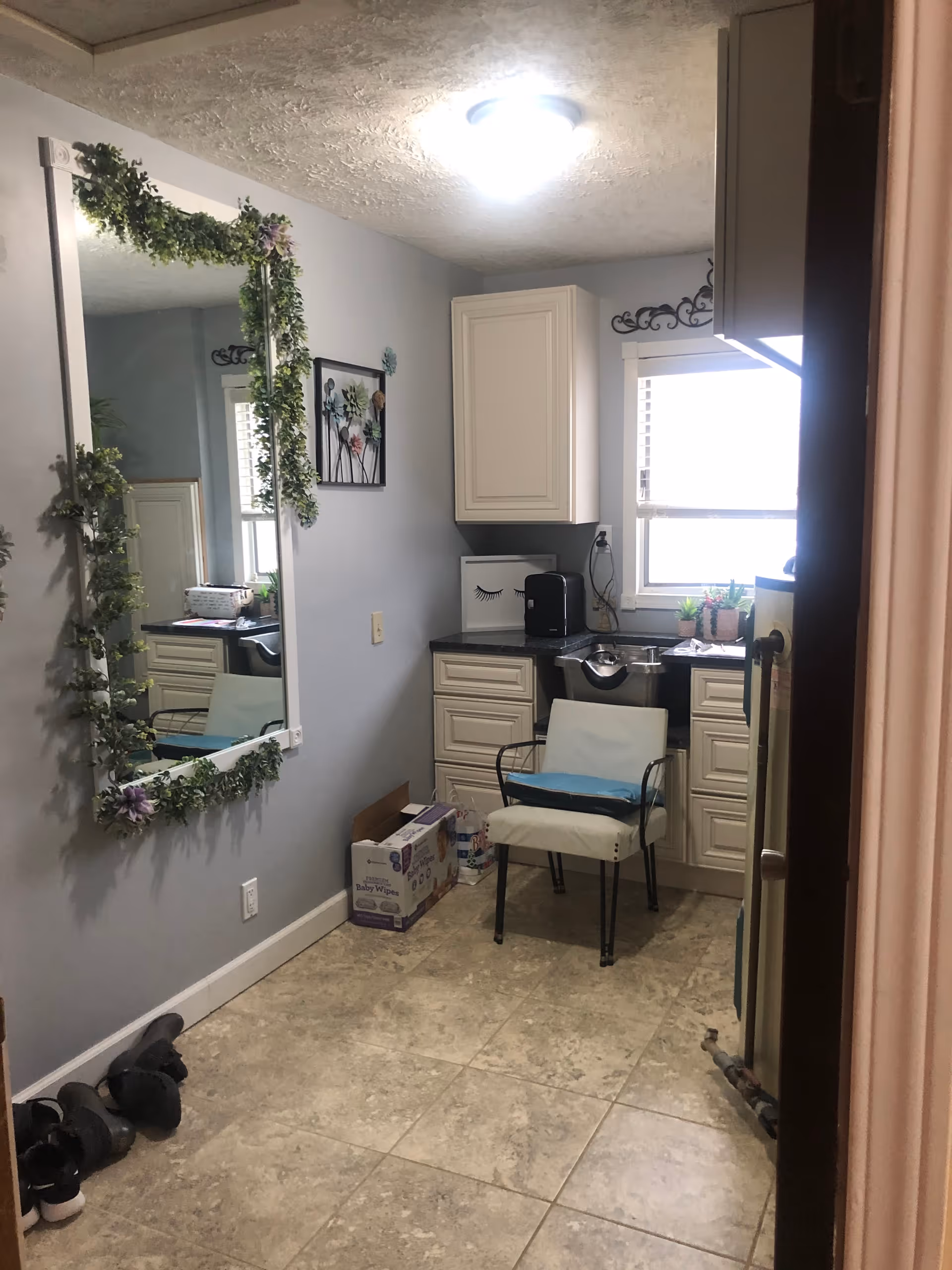 A small room with light gray walls and tiled floor featuring a white cushioned chair with black metal arms and legs, a countertop with white cabinets above and below, a window with blinds, a large wall mirror decorated with green garland, a wall art piece with flowers, and some boxes and shoes on the floor.