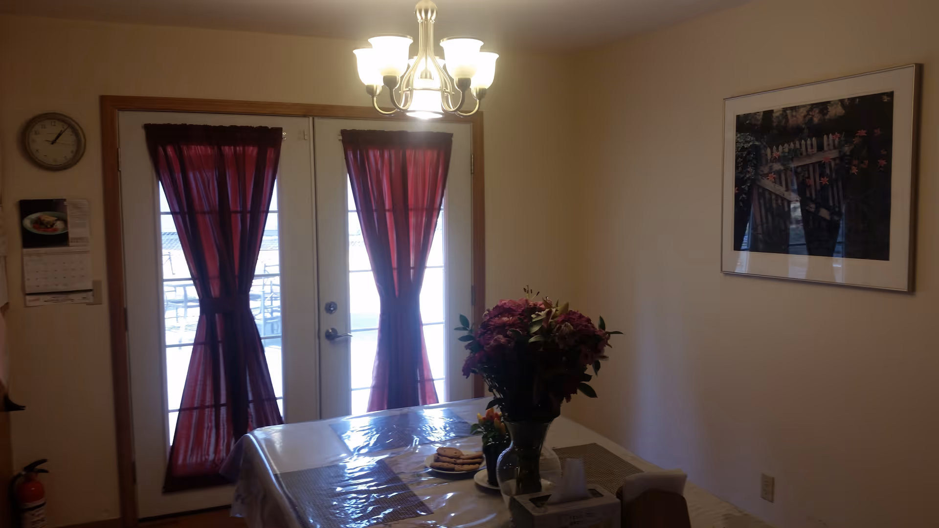 Dining area with a table covered by a plastic tablecloth, a vase with flowers, and a plate with cookies. Behind the table are double glass doors with red curtains tied in the middle. A chandelier with five lights hangs from the ceiling. On the wall to the right is a framed picture, and on the left wall is a clock and a calendar.