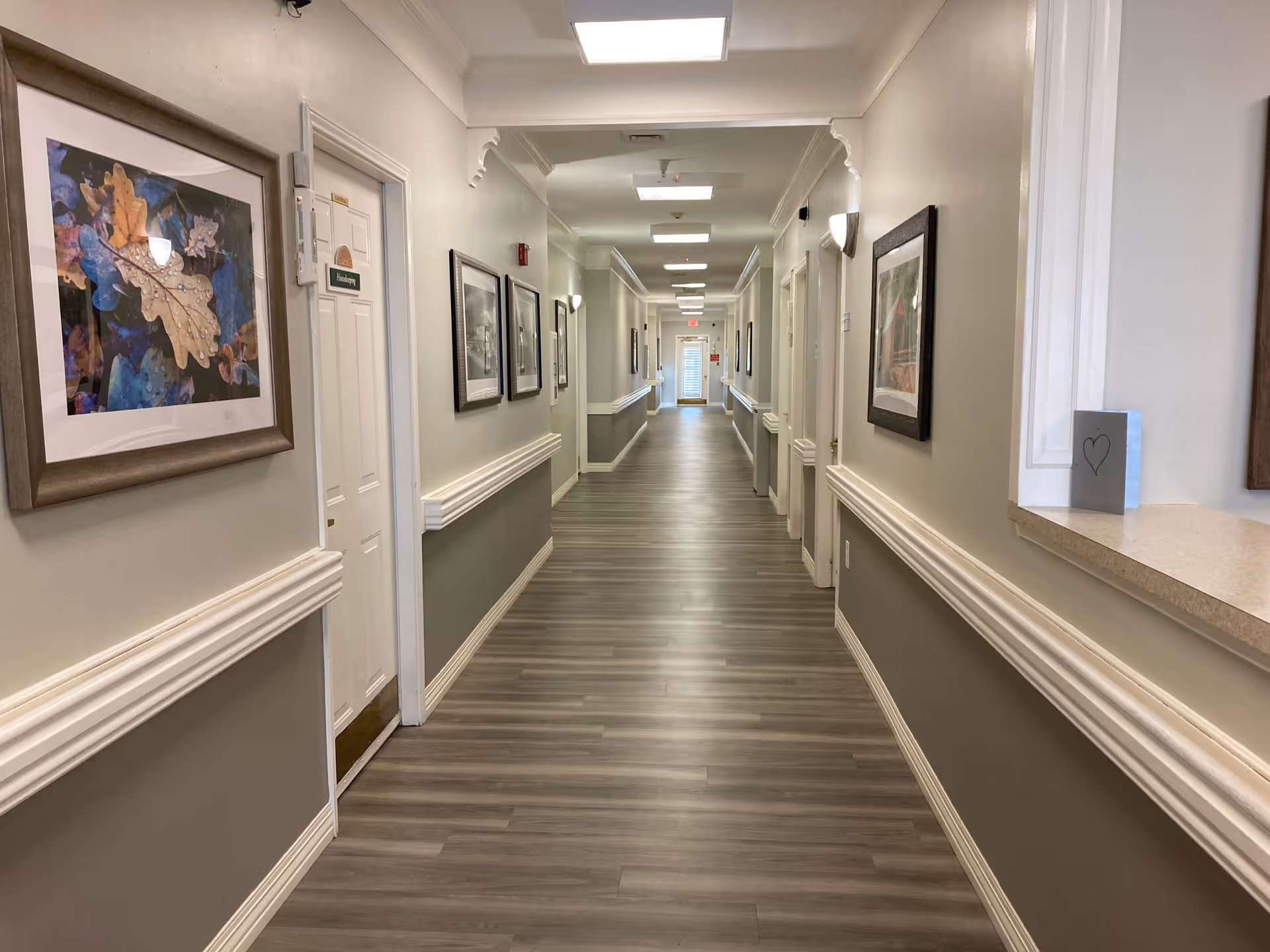 Long, well-lit interior hallway of a senior living facility with framed artwork and handrails along both walls.