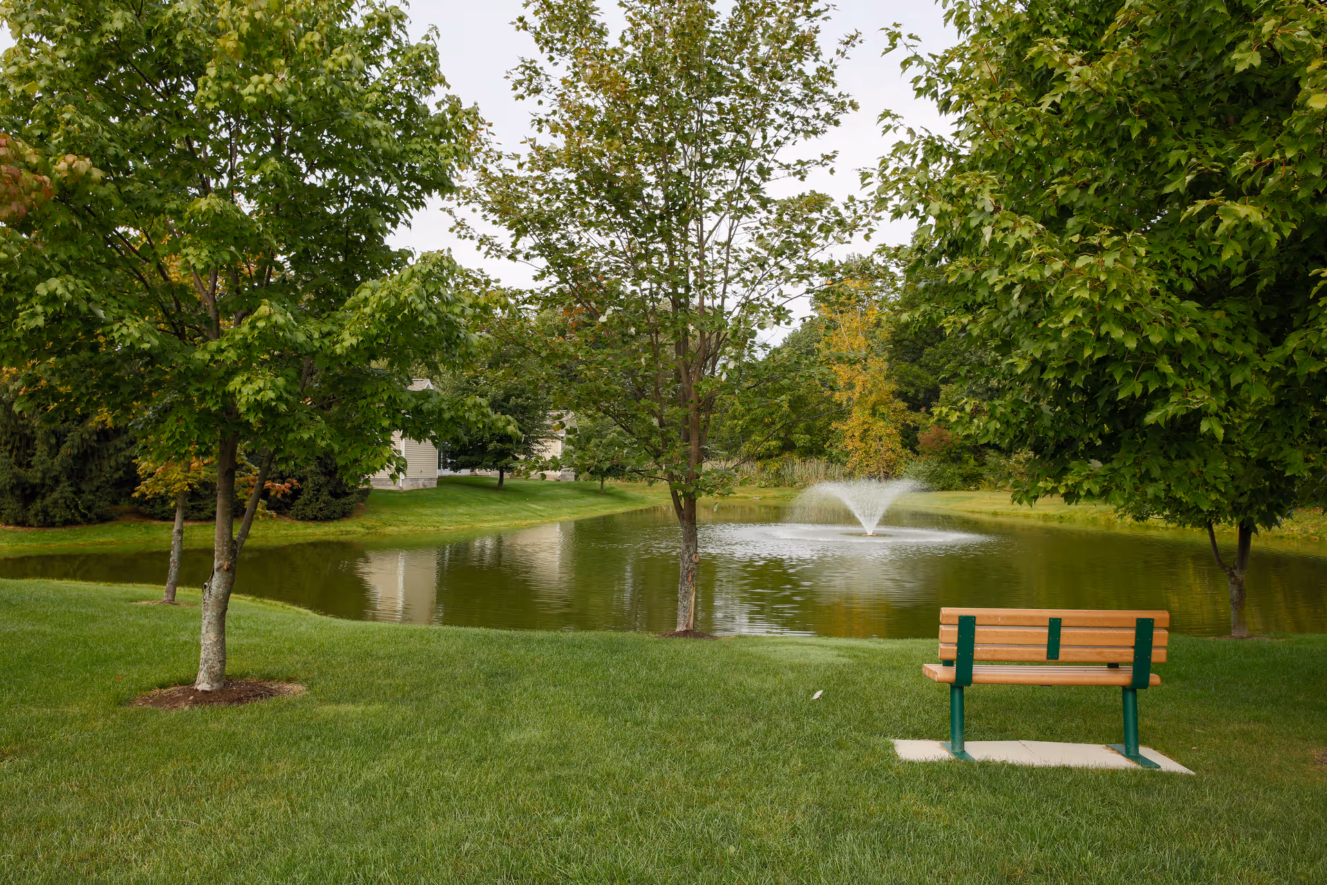 A peaceful outdoor scene at The Village Townhomes featuring a small pond with a water fountain in the center, surrounded by green grass and several leafy trees. A wooden bench with green metal supports is positioned on the grass facing the pond.