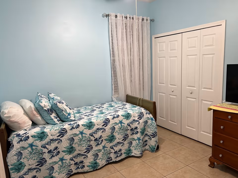 Small bedroom with a single bed covered in teal and blue patterned bedding, white bi-fold closet doors, a dresser and a TV.