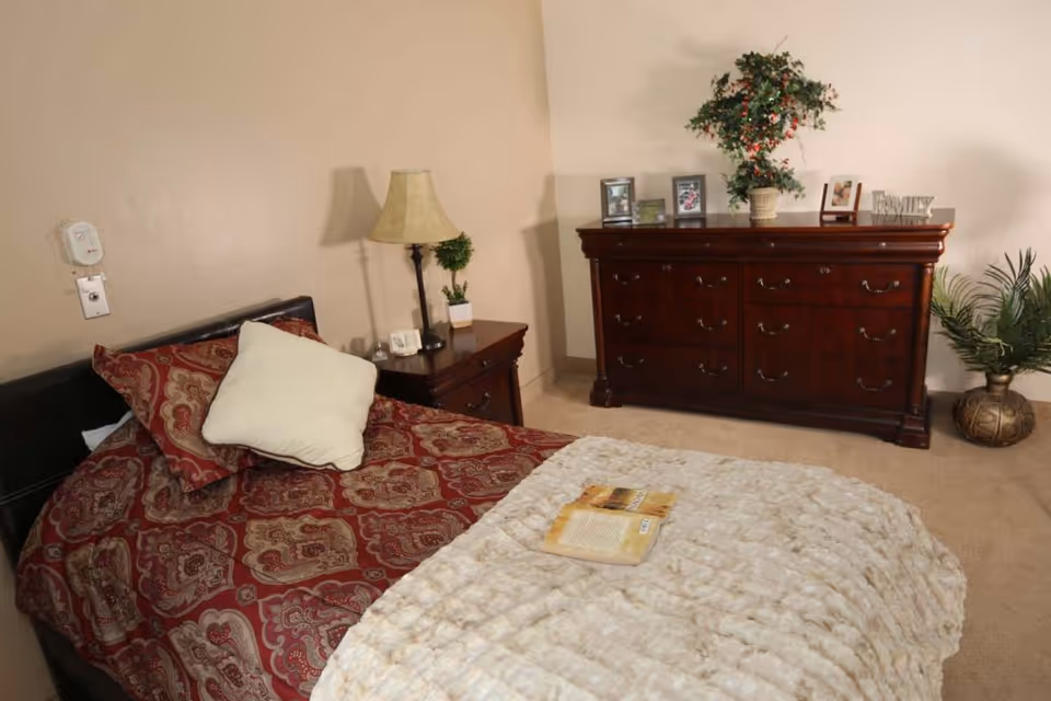 A furnished bedroom with a bed and pillows, a nightstand with a lamp, and a wooden dresser topped with plants and framed photos.