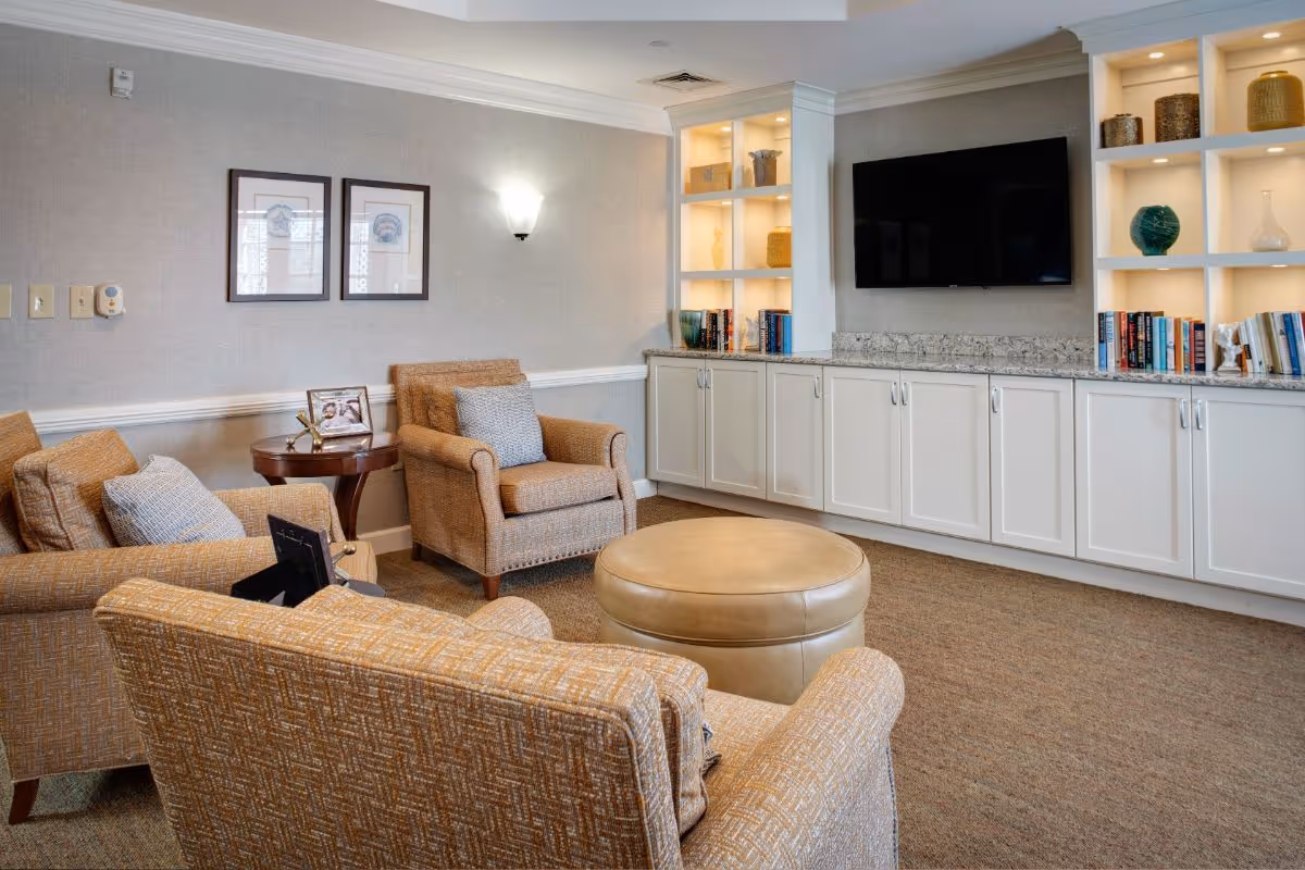 Comfortable common living room with upholstered armchairs, a round ottoman, built-in cabinets and shelves, and a wall-mounted TV.