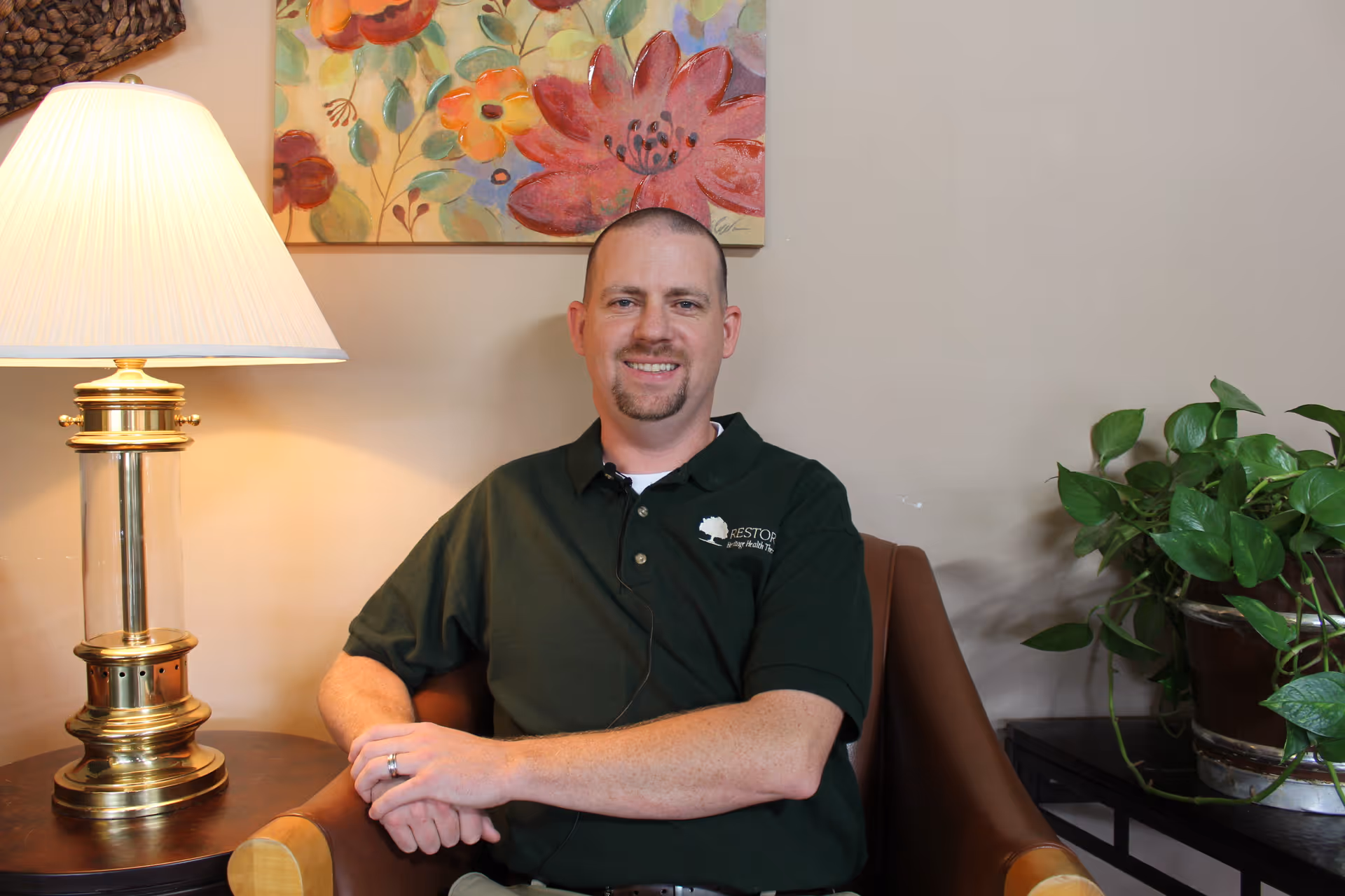 A man with a short beard and mustache is sitting in a brown armchair in a cozy room. He is wearing a dark green polo shirt with a logo on it. To his left is a table with a brass and glass lamp, and behind him is a colorful floral painting on the wall. To his right, there is a green leafy plant on a small table.
