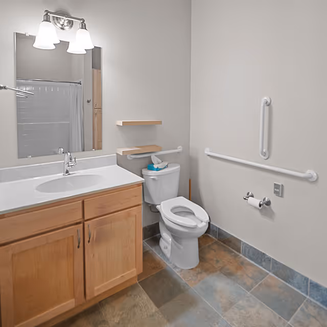 Accessible bathroom with a sink vanity and mirror on the left, a toilet with grab bars, and tiled floor.